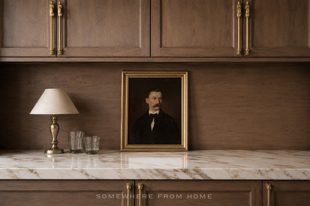 A framed vintage portrait of a man with a mustache in a dark suit, displayed on a white marble countertop against a dark wood cabinet wall, next to a table lamp and two glasses.