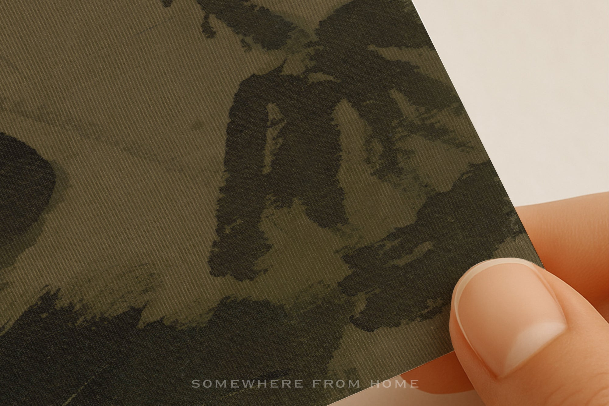 A close-up of a fabric or paper sample with a dark abstract pattern, held by a person's fingers against a plain light background. The text 'SOMEWHERE FROM HOME' appears near the bottom.