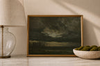 A framed landscape painting with a dark stormy sky and cypress trees, displayed on a marble surface next to a table lamp and a bowl of green moss.