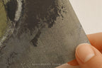 A close-up of a textured fabric or canvas print being held by a hand, showing a dark abstract painted pattern in charcoal, gray, and black tones.