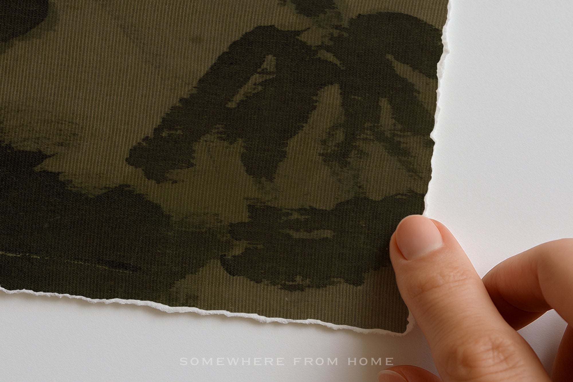 A close-up of a hand holding the edge of a piece of fabric with a dark olive green and black camouflage pattern, set against a plain white background.