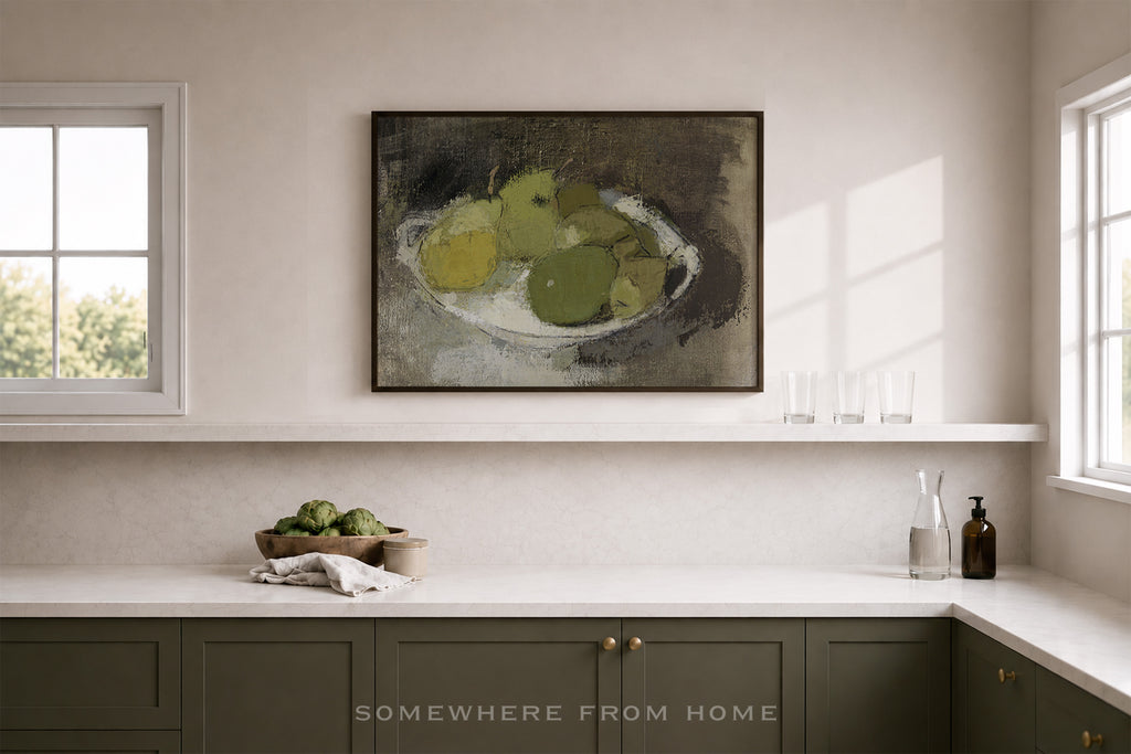 A framed still life painting of green and yellow pears on a shelf above a kitchen counter in a bright, modern kitchen. The cabinets are a muted sage green, and the walls are a warm off-white.