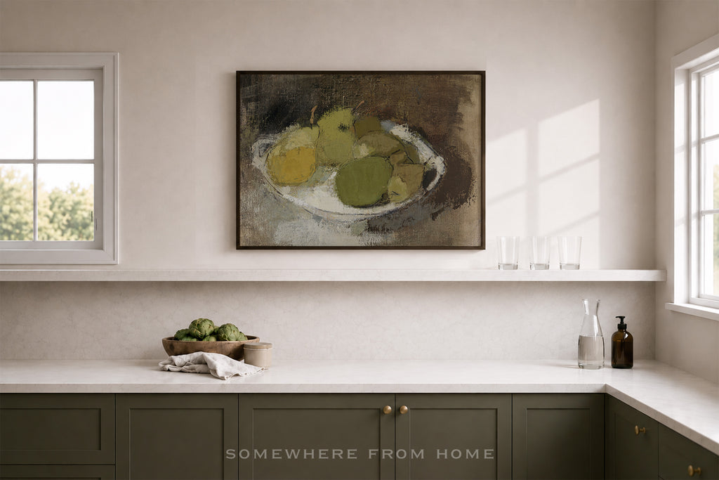 A framed still life painting of green and yellow pears on a shelf above a kitchen counter in a bright, modern kitchen. The cabinets are a muted sage green, and the walls are a warm off-white.