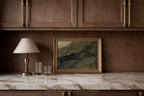A framed abstract landscape painting with muted green, gray, and black tones in a gold frame, displayed on a white marble countertop against a dark wood cabinet wall, next to a table lamp and two glasses.