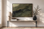A framed abstract landscape art print with green, olive, and dark gray brushstrokes hangs on a beige wall above a console table styled with a lamp, books, and a black vase with branches.