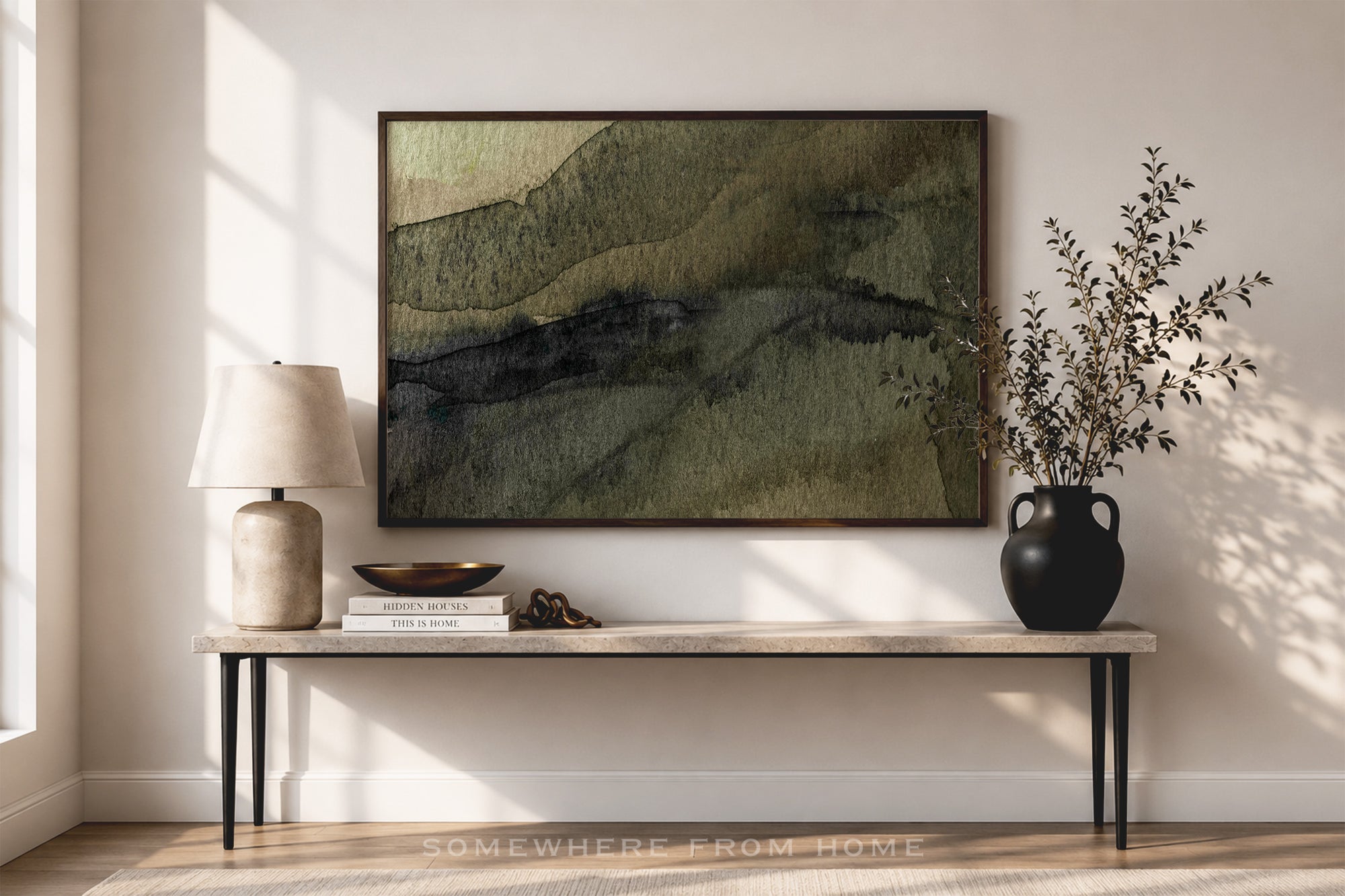 A framed abstract landscape art print with green, olive, and dark gray brushstrokes hangs on a beige wall above a console table styled with a lamp, books, and a black vase with branches.