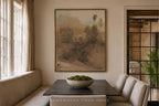A framed landscape art print hangs on a beige wall above a dark wood dining table with upholstered chairs in a softly lit room. The artwork depicts muted earthy hills and trees in brown, beige, and gray tones.