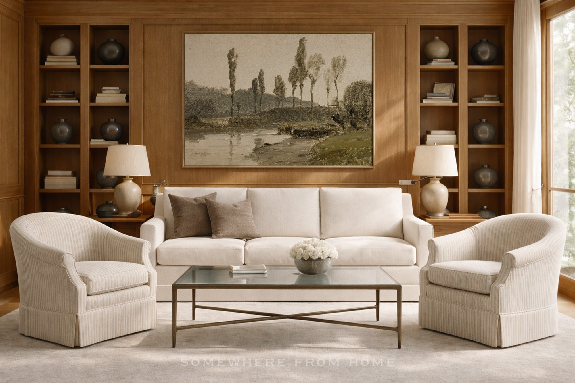 A styled living room featuring a cream-colored three-seater sofa flanked by two matching armchairs, a glass-top coffee table, and a large framed landscape painting on a wood-paneled wall.