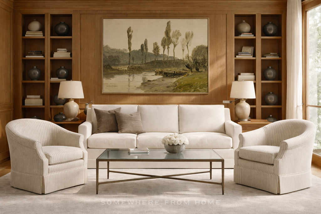 A styled living room featuring a cream-colored three-seater sofa flanked by two matching armchairs, a glass-top coffee table, and a large framed landscape painting on a wood-paneled wall.