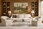 A styled living room featuring a cream-colored three-seater sofa flanked by two matching armchairs, a glass-top coffee table, and a large framed landscape painting on a wood-paneled wall.