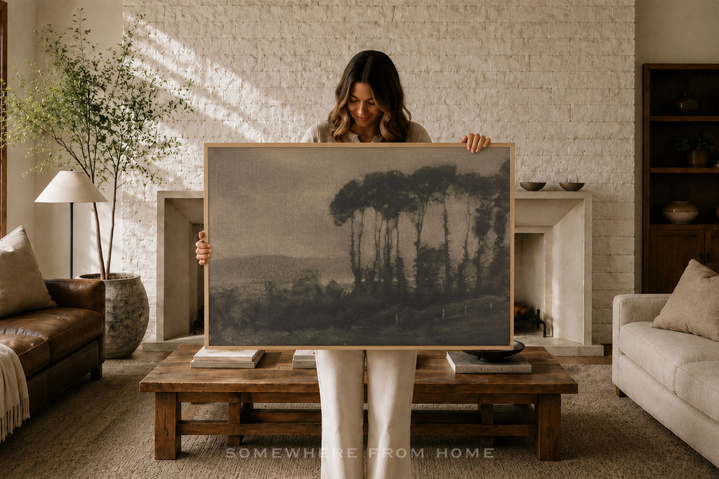 A woman stands in a cozy living room holding up a large framed landscape art print. The artwork depicts a moody, dark forest scene with tall pine-like trees under a muted gray sky.