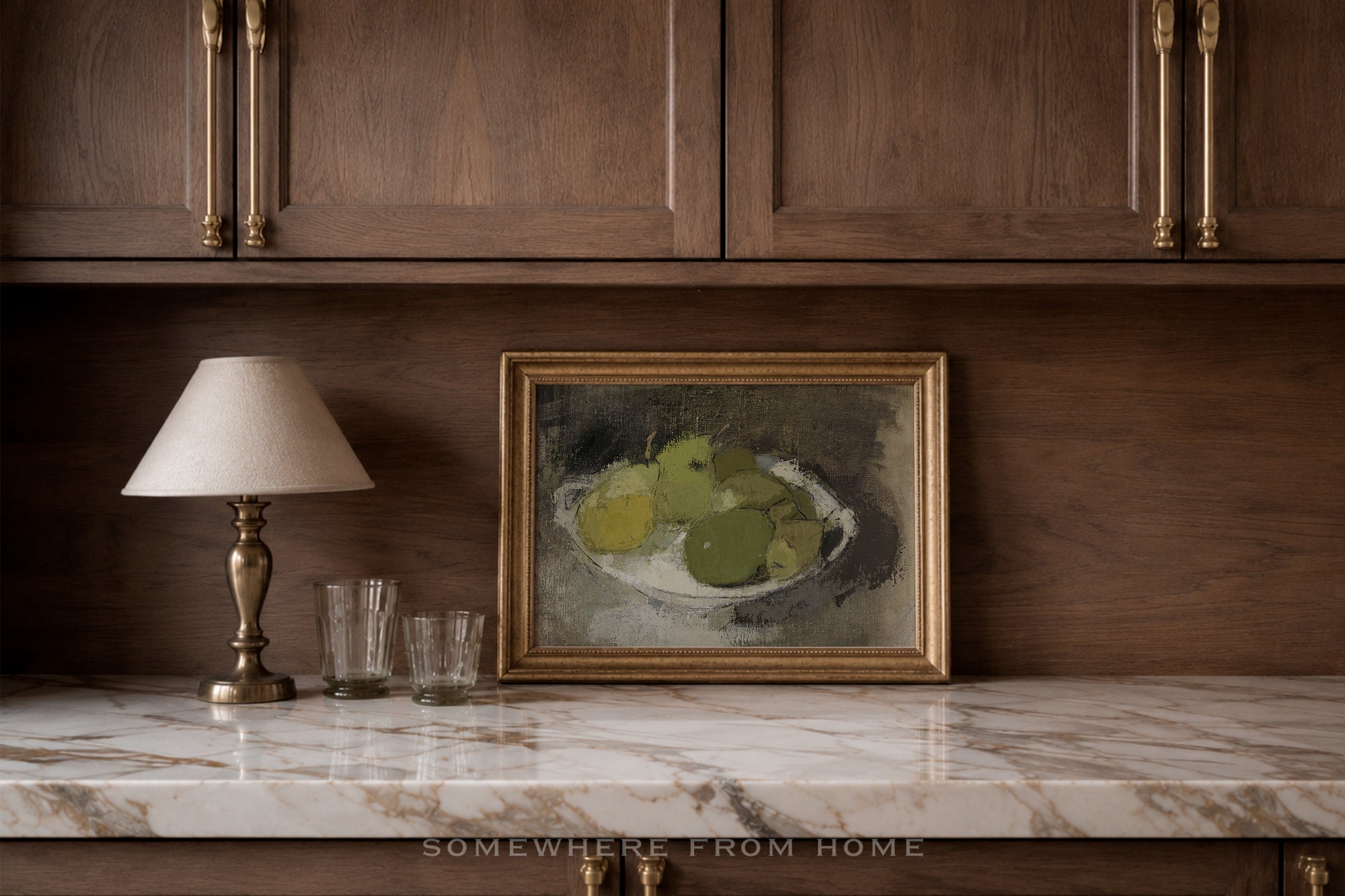 A framed still life painting of green apples in a bowl, displayed on a marble countertop against a dark wood cabinet wall, next to a table lamp and two glasses.