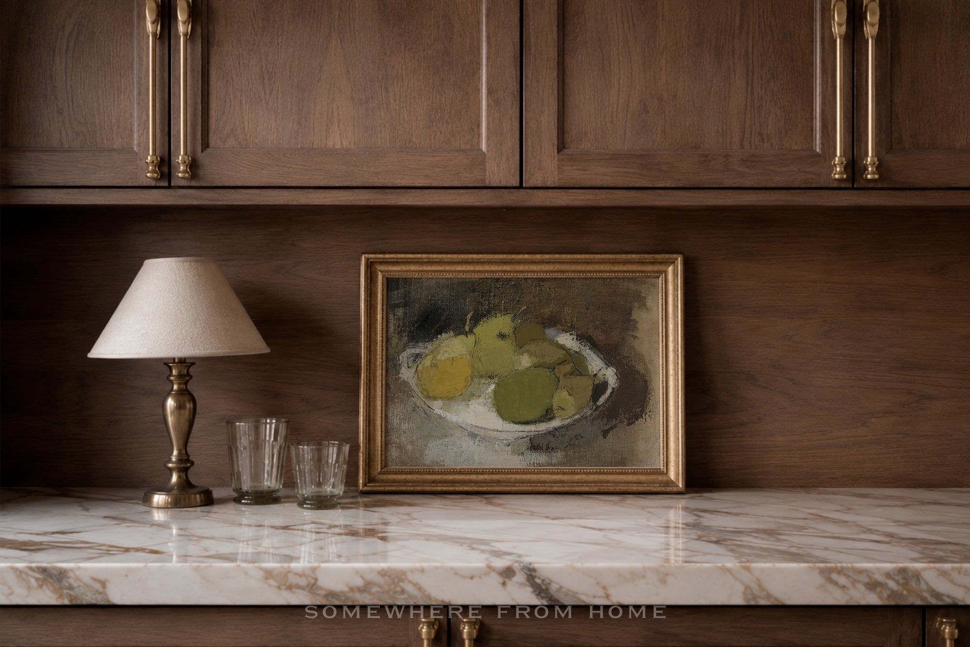 A framed still life painting of green and yellow pears in a white bowl, displayed on a marble countertop against a dark wood cabinet wall, next to a table lamp and two glasses.