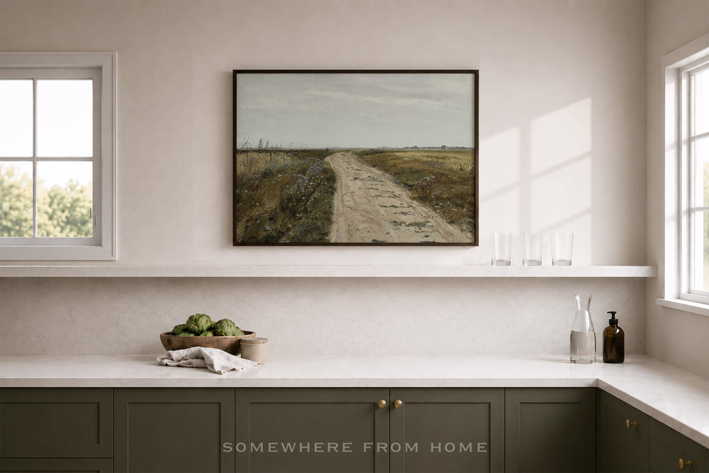 A framed landscape painting of a dirt road through a field hangs on a beige wall above a white shelf in a modern kitchen setting. The kitchen features dark green cabinets, a white countertop, and two windows.