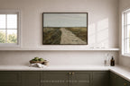 A framed landscape painting of a dirt road through a field hangs on a beige wall above a white shelf in a modern kitchen setting. The kitchen features dark green cabinets, a white countertop, and two windows.