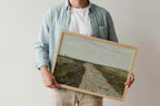 A person holding a framed landscape painting of a dirt road through a grassy field under a cloudy sky. The person is wearing a light blue denim shirt over a white t-shirt.