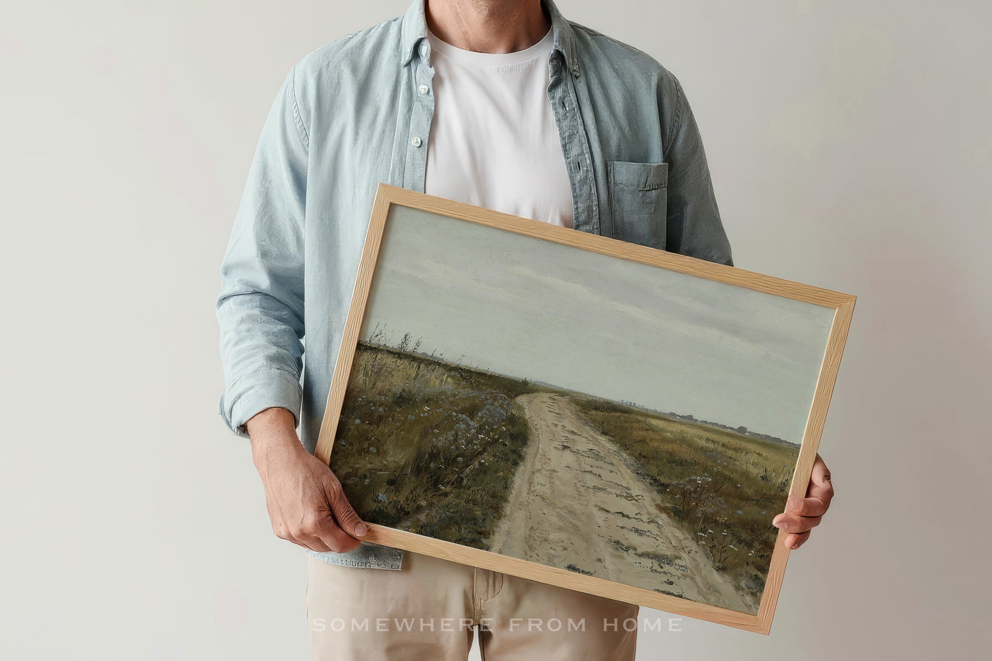 A person holding a framed landscape painting of a dirt road through a grassy field under a cloudy sky. The person is wearing a light blue denim shirt over a white t-shirt.