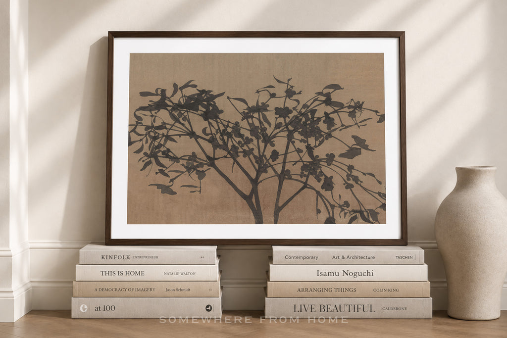 A framed art print featuring a black ink-style tree silhouette on a warm beige background, leaning against a cream wall above a stack of books, with a ceramic vase to the right.