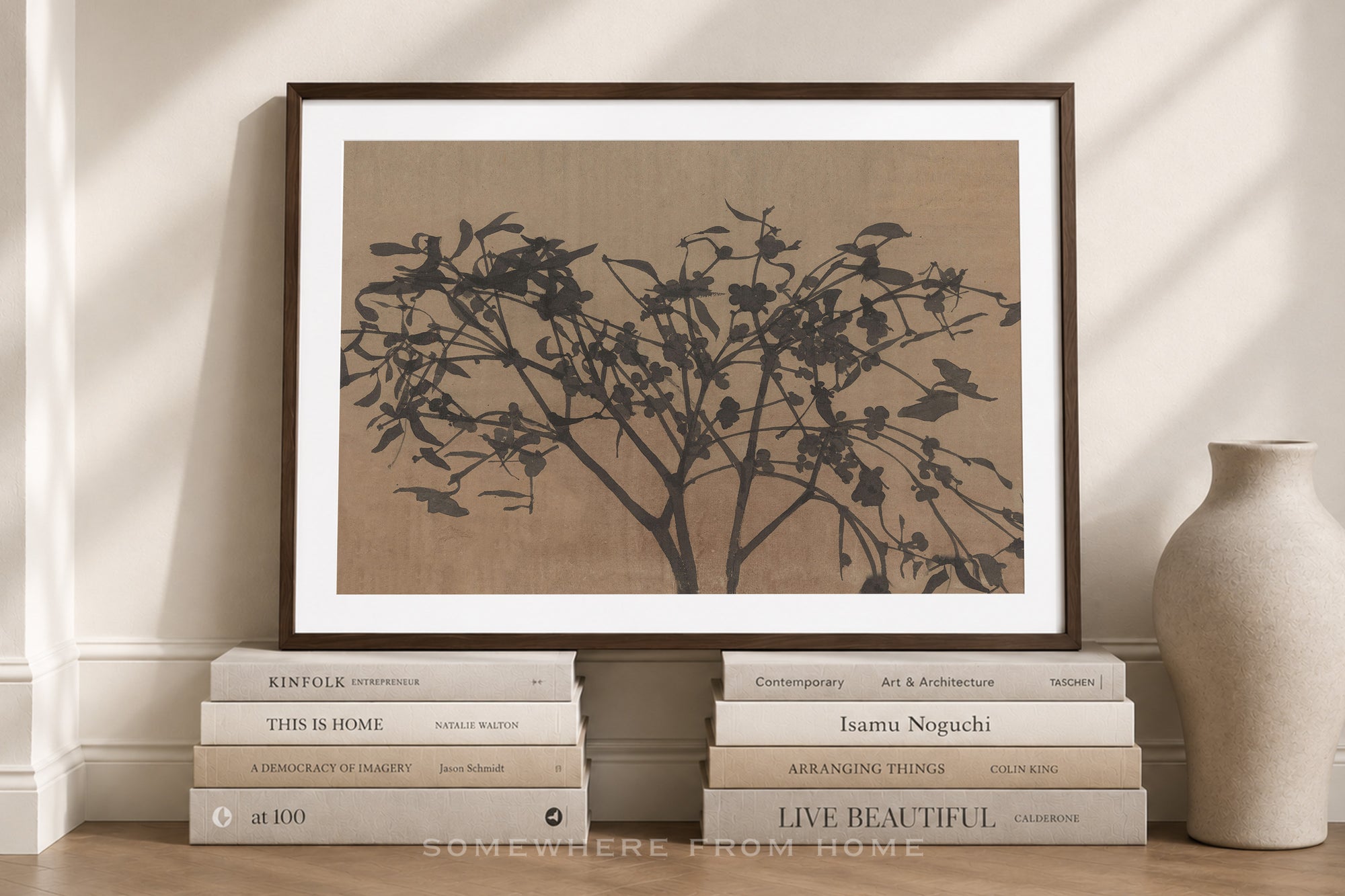 A framed art print featuring a black ink-style tree silhouette on a warm beige background, leaning against a cream wall above a stack of books, with a ceramic vase to the right.