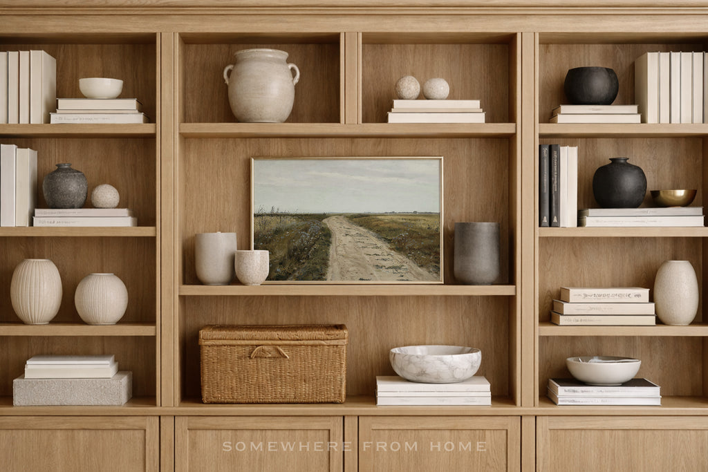A styled interior scene showing a large light wood bookcase filled with books, ceramic vases, bowls, and a framed landscape painting. The text 'SOMEWHERE FROM HOME' appears at the bottom.
