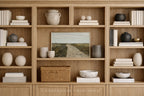 A styled interior scene showing a large light wood bookcase filled with books, ceramic vases, bowls, and a framed landscape painting. The text 'SOMEWHERE FROM HOME' appears at the bottom.