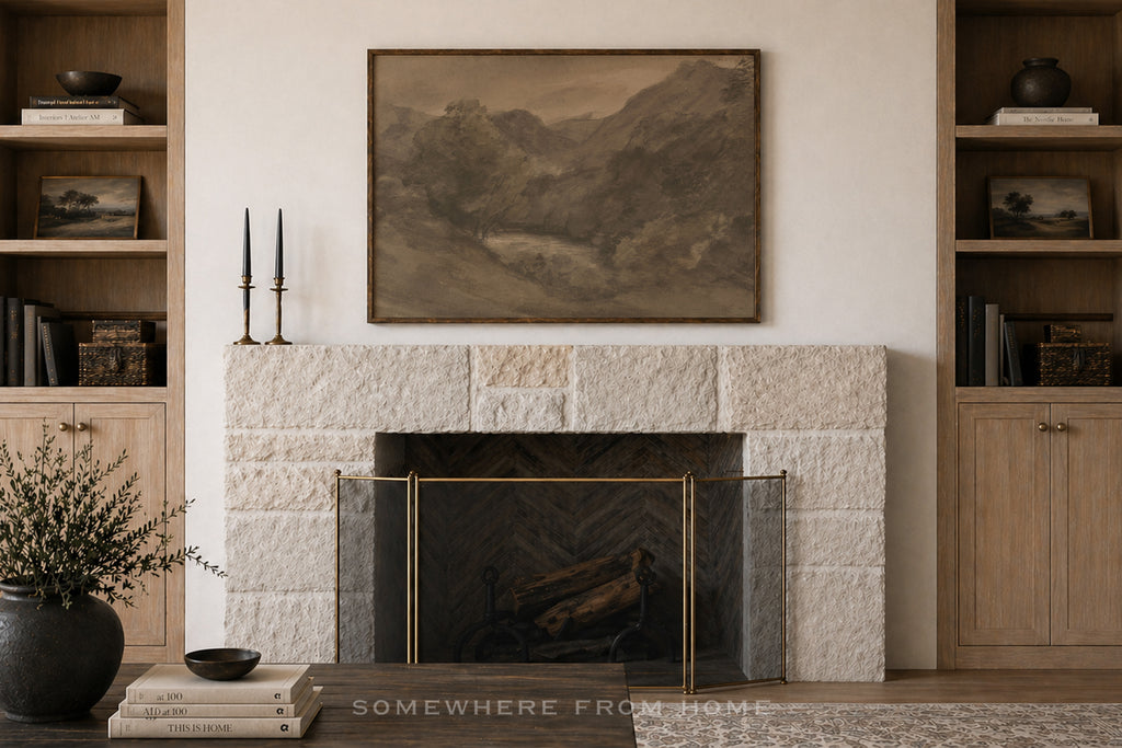 A framed landscape art print in muted earth tones hangs above a stone fireplace in a styled room with wooden shelves, candles, books, and a vase.