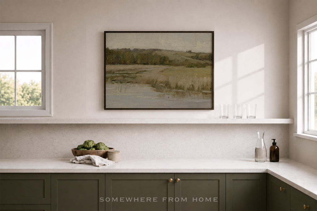 A framed landscape art print hangs on a light beige wall above a white shelf in a modern kitchen setting. The print depicts a muted, earthy scene of rolling hills and a river, and the kitchen features dark green cabinets, a white countertop, and two windows.