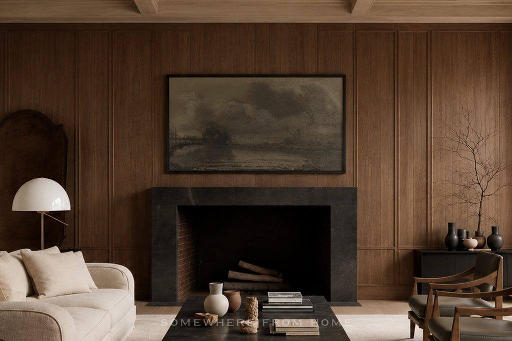 A framed landscape art print in muted gray and brown tones hangs above a dark fireplace in a warm wood-paneled living room with a cream sofa, black coffee table, and sideboard.