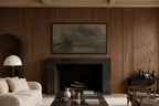 A framed landscape art print in muted gray and brown tones hangs above a dark fireplace in a warm wood-paneled living room with a cream sofa, black coffee table, and sideboard.