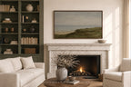 A framed landscape painting of green dunes and a beach hangs above a white marble fireplace in a styled living room with a cream sofa, armchair, shelving, and a lit fire.