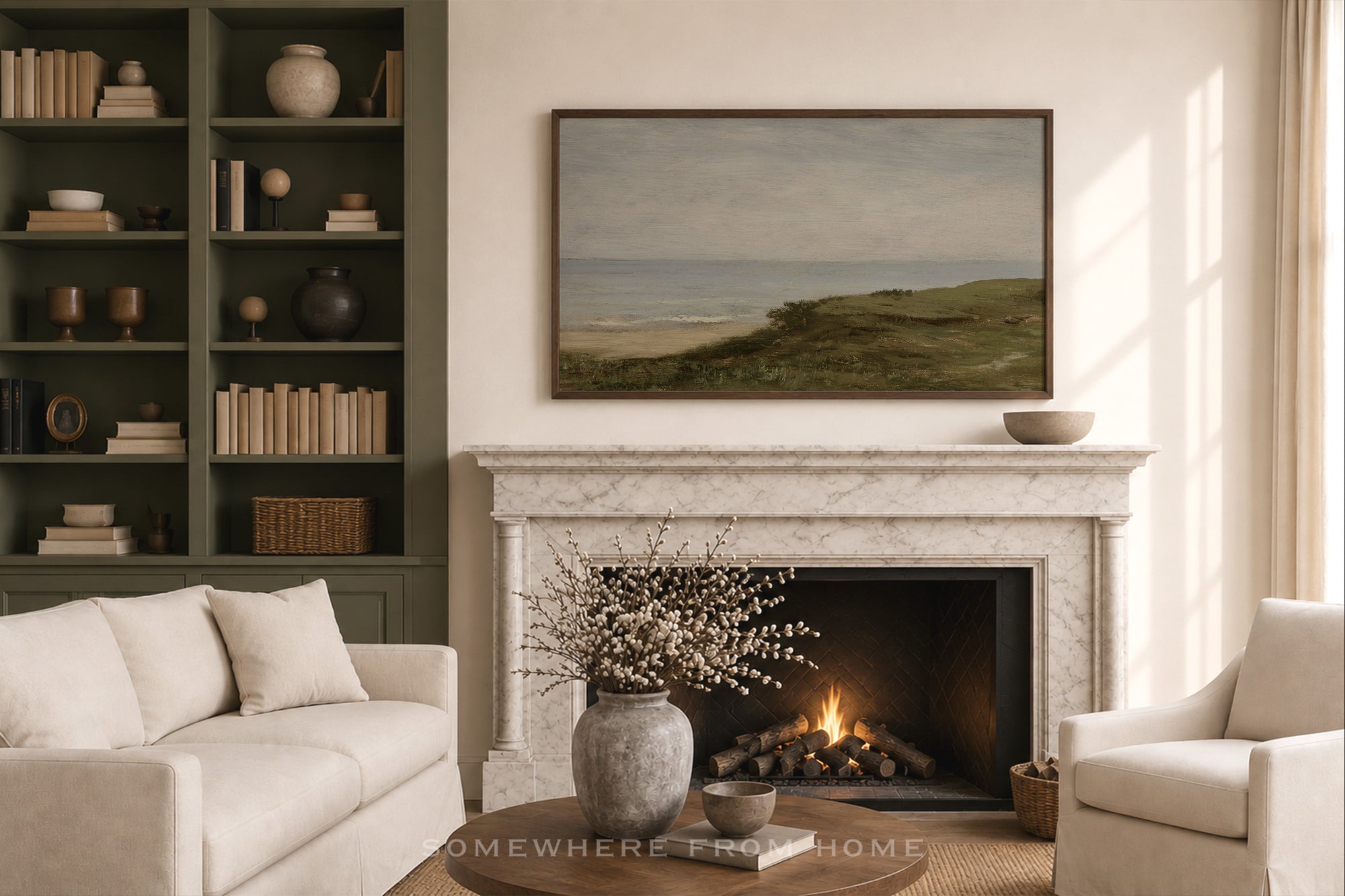 A framed landscape painting of green dunes and a beach hangs above a white marble fireplace in a styled living room with a cream sofa, armchair, shelving, and a lit fire.