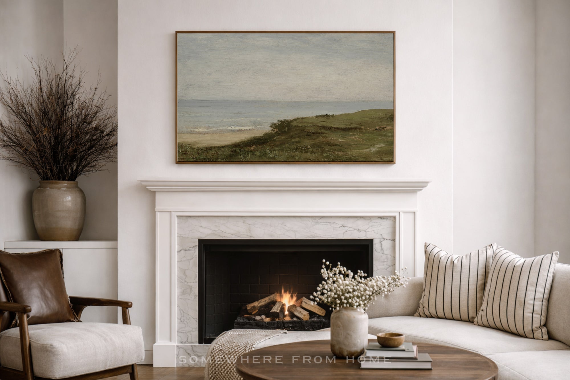 A framed landscape painting of a grassy dune field leading to the ocean, displayed in a living room above a white marble fireplace mantel.