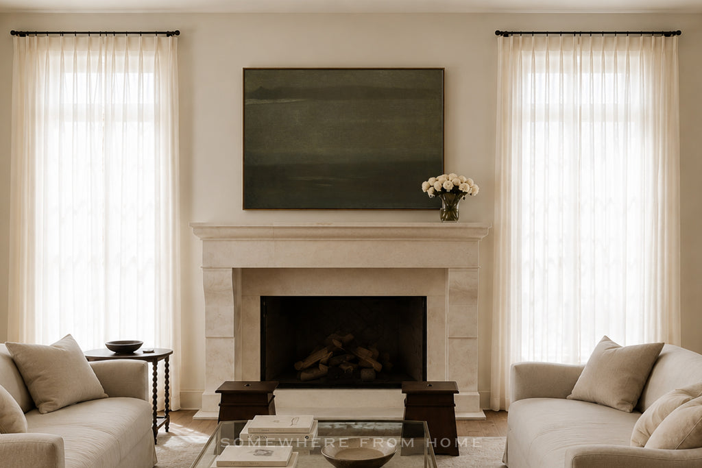 A styled interior room featuring a white stone fireplace with a dark green rectangular wall art piece above it, flanked by two tall windows with sheer white curtains. Light beige sofas sit on either side, with a glass coffee table in front.