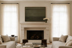 A styled interior room featuring a white stone fireplace with a dark green rectangular wall art piece above it, flanked by two tall windows with sheer white curtains. Light beige sofas sit on either side, with a glass coffee table in front.