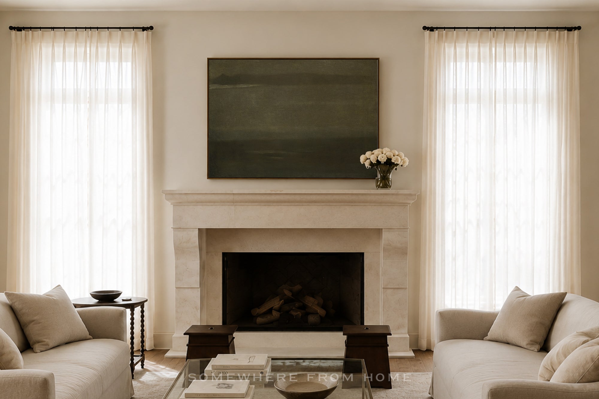 A styled interior room featuring a white stone fireplace with a dark green rectangular wall art piece above it, flanked by two tall windows with sheer white curtains. Light beige sofas sit on either side, with a glass coffee table in front.