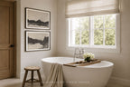 A modern freestanding white bathtub in a bright, neutral-toned bathroom with a window, beige roman shade, wooden stool, and two framed black-and-white landscape prints on the wall. A wooden tray with white flowers rests across the tub, and a towel is draped over the edge.