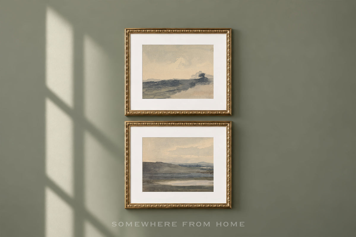 Two framed landscape art prints with gold frames and white mats, hanging vertically on a muted sage green wall with soft window shadow cast across it. The text 'SOMEWHERE FROM HOME' appears below the frames.