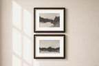 Two framed black and white landscape art prints with gold inner trim and black outer frames, hanging vertically on a beige wall with soft window shadow light.