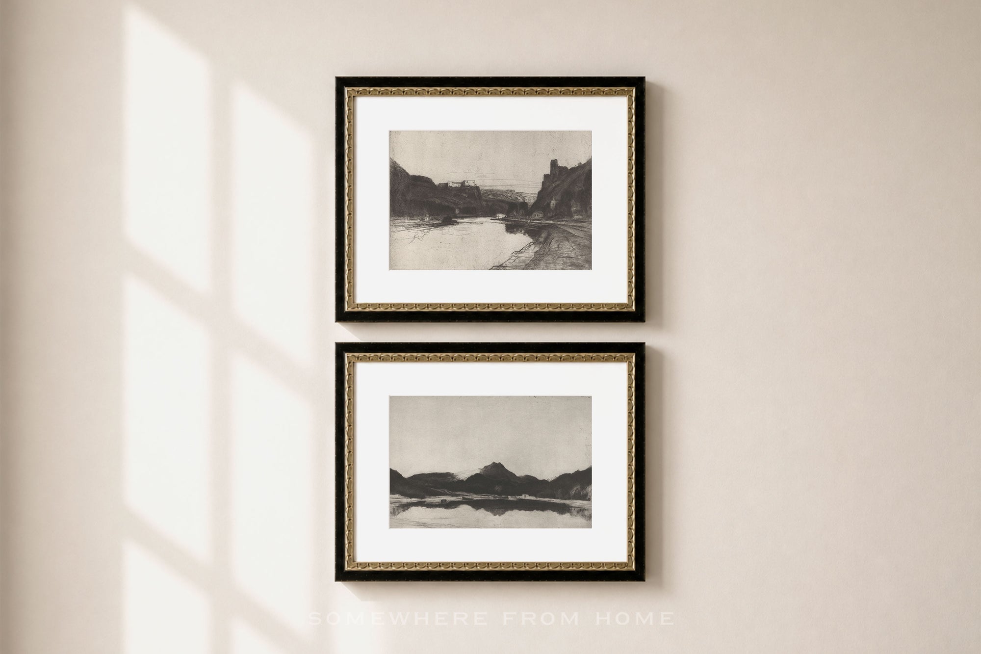 Two framed black and white landscape art prints with gold inner trim and black outer frames, hanging vertically on a beige wall with soft window shadow light.