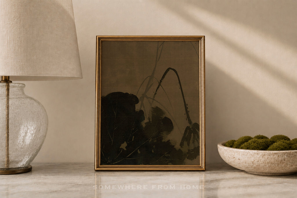 A framed art print featuring a dark, moody abstract landscape with black and brown tones, styled on a marble surface next to a table lamp and a bowl of green moss.