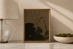 A framed art print featuring a dark, moody abstract landscape with black and brown tones, styled on a marble surface next to a table lamp and a bowl of green moss.