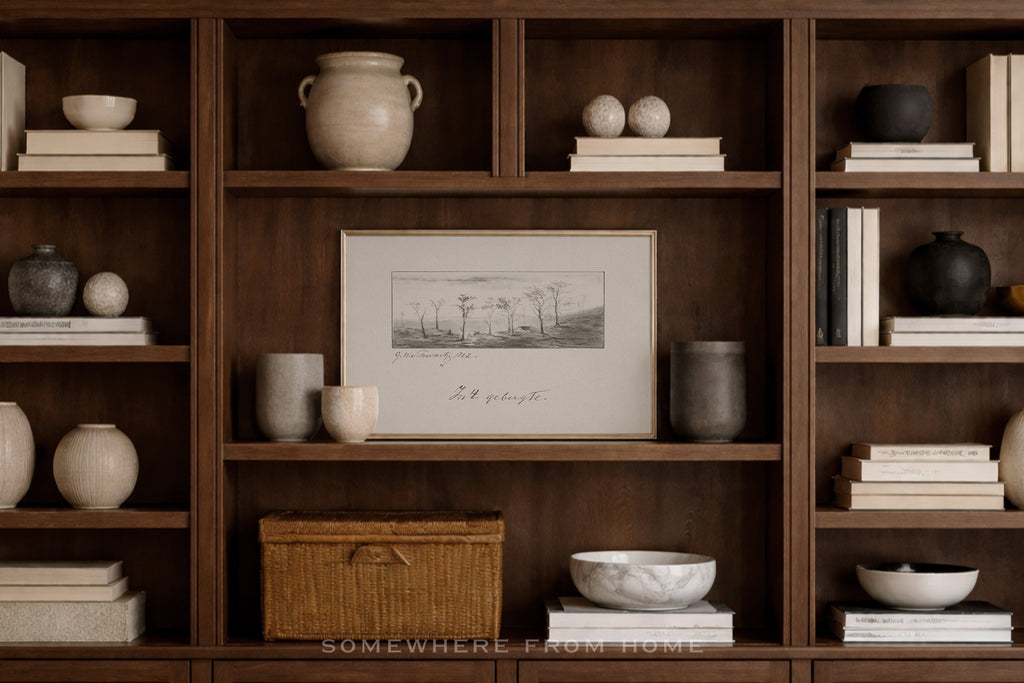 A framed black-and-white landscape sketch hangs on a dark wood bookshelf styled with ceramic vases, books, bowls, and a wicker box in a warm, neutral interior setting.