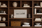 A framed black-and-white landscape sketch hangs on a dark wood bookshelf styled with ceramic vases, books, bowls, and a wicker box in a warm, neutral interior setting.