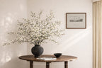 Decorative vase with white flowers on a wooden table against a neutral wall with a framed picture.