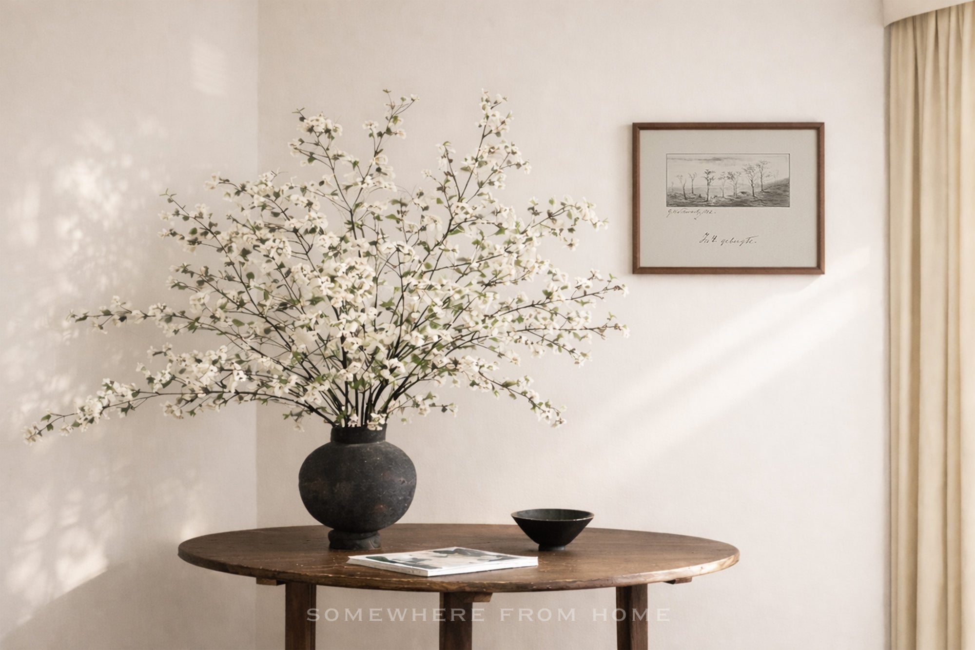 Decorative vase with white flowers on a wooden table against a neutral wall with a framed picture.