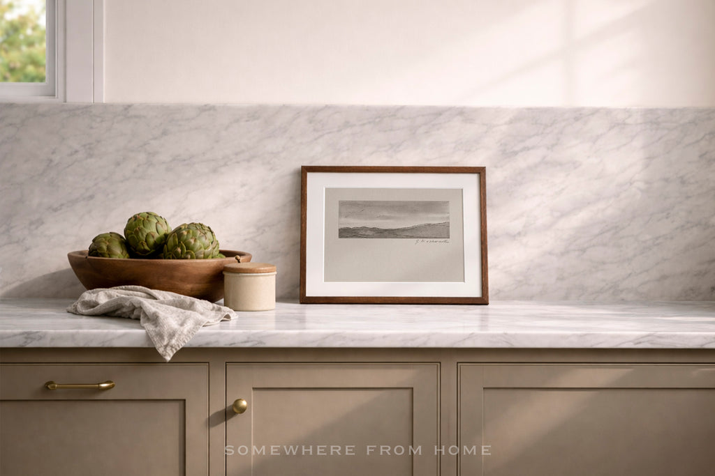 A framed black and white landscape print with a medium brown wood frame sits on a white marble kitchen counter against a matching marble backsplash. The scene includes a wooden bowl of artichokes, a small jar, and a folded cloth, with a window light casting soft shadows.