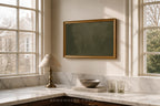 A framed dark green canvas print with a gold frame hanging on a beige wall above a marble kitchen counter with a lamp, bowl, and glasses.