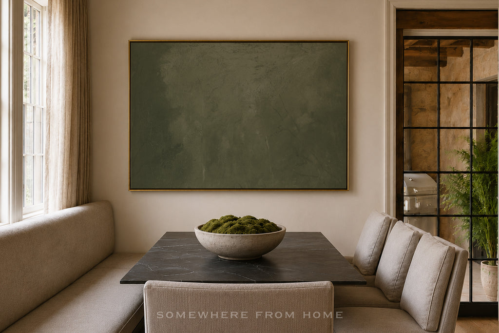 A large framed abstract wall art piece in muted olive green hangs above a dining nook with beige upholstered seating and a dark table. The room has warm neutral walls, a window with curtains, and a glass-paneled door with a plant visible outside.