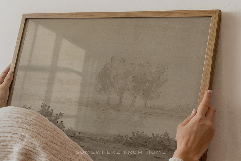 A framed art print featuring a muted landscape with three trees, held by hands against a light wall. The print has a warm beige background with a gray-toned illustration, enclosed in a light wood frame.