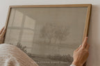A framed art print featuring a muted landscape with three trees, held by hands against a light wall. The print has a warm beige background with a gray-toned illustration, enclosed in a light wood frame.